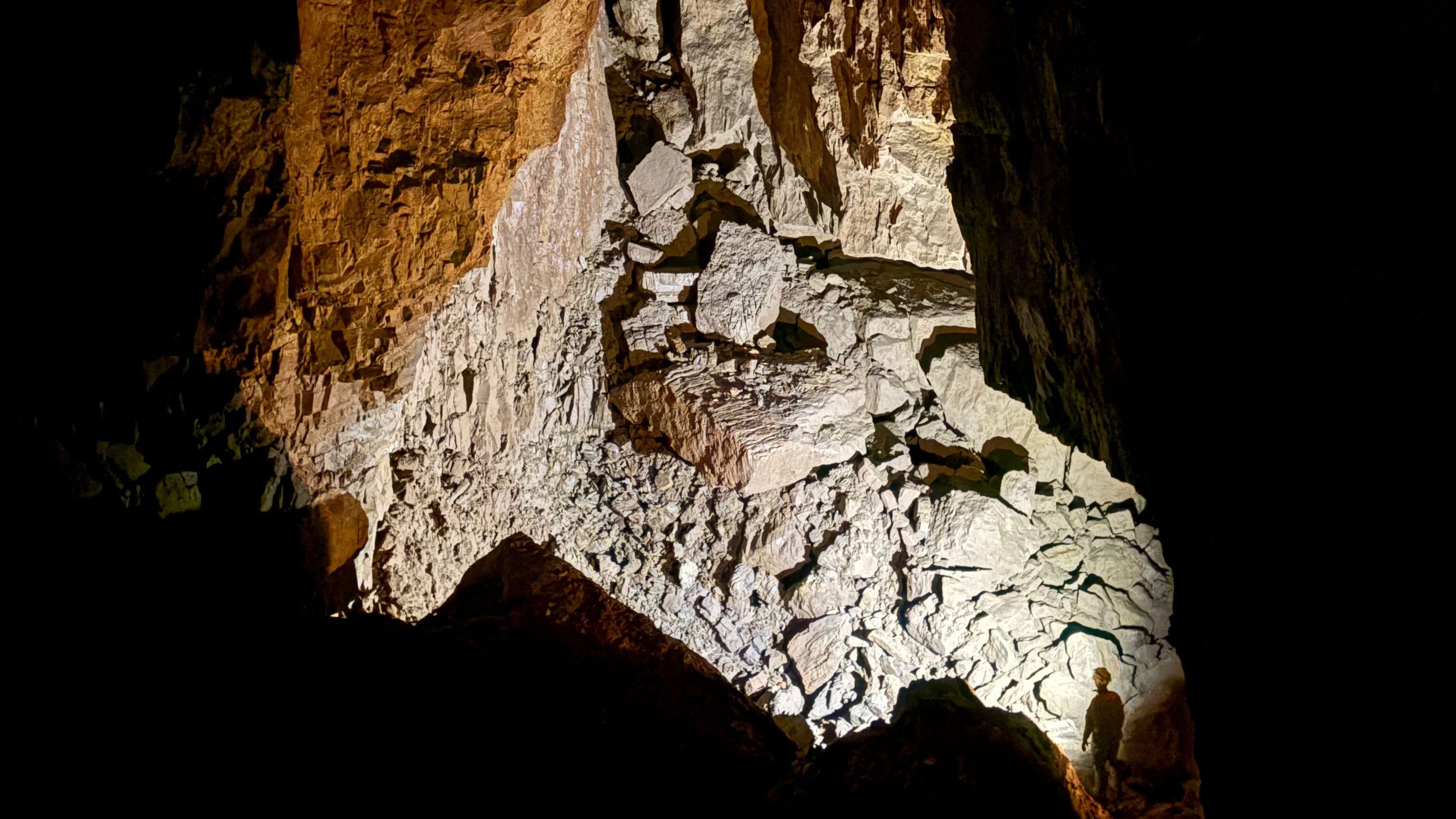 A lone caver dwarfed by a lit wall of boulders in a huge dark chamber
